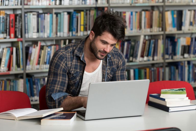 Student w okularach pisze pracę inżynierską na laptopie otoczony schematami i książkami inżynierskimi
