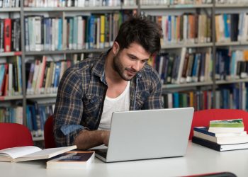 Student w okularach pisze pracę inżynierską na laptopie otoczony schematami i książkami inżynierskimi